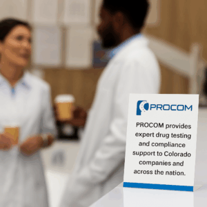 Two healthcare professionals in white coats talk casually in a modern clinic while a branded PROCOM informational brochure is displayed on the reception counter in focus.