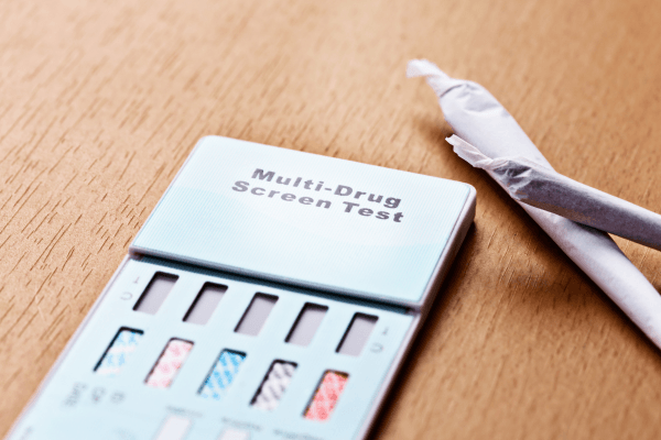 A multi-drug screen test device panel resting on a wooden surface next to hand-rolled joints, representing the point of a positive result notification.
